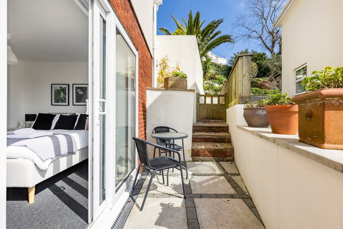 A private patio area is visible, featuring a small round table and two chairs. Steps lead up to a landscaped path with potted plants. Sunlight illuminates the area, and a spacious room with a light-colored bed can be seen in the background through glass doors.