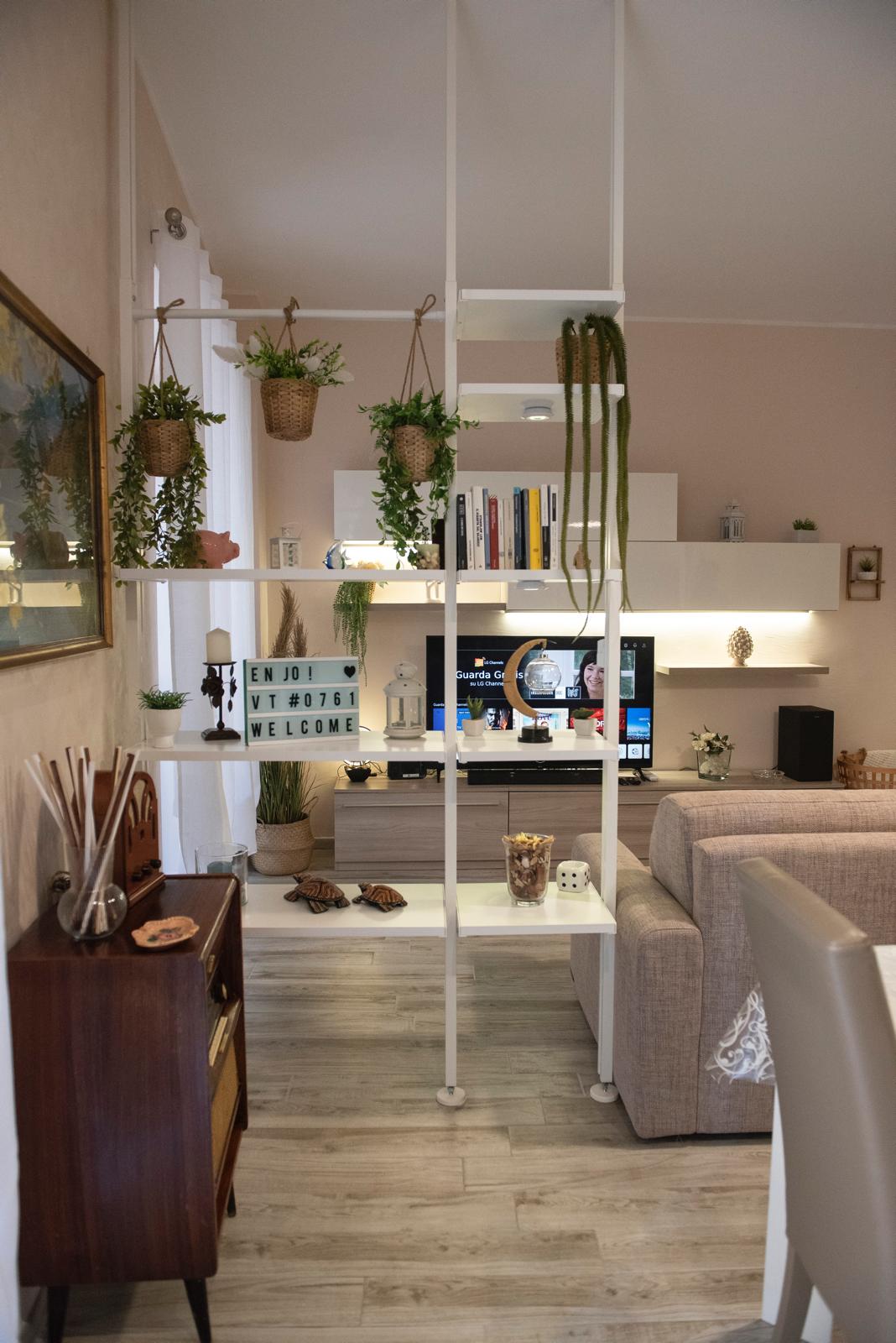 A stylish open-space living area features a neutral color palette with indoor plants displayed on shelves. A Smart TV is positioned on a minimalist media unit, while a comfortable sofa invites relaxation. A decorative vintage cabinet adds charm to the room.