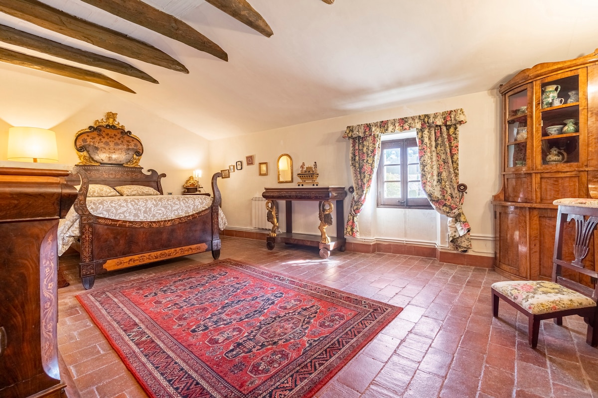 A spacious bedroom features wooden beams on the ceiling and terracotta tiled flooring. A large antique bed with floral upholstery is centered against the wall, accompanied by a wooden dresser and a decorative side table. Natural light filters through a window, framed by floral curtains.