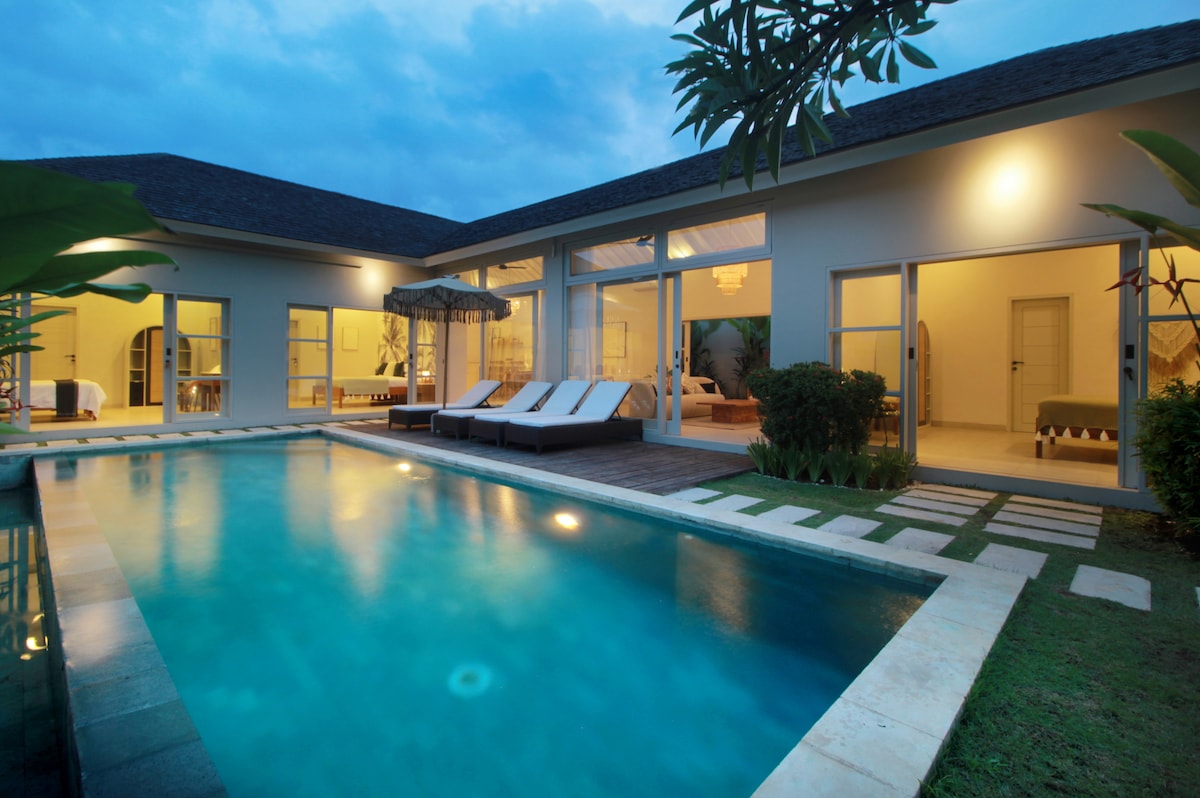 An inviting villa exterior is showcased, featuring a private pool with calm blue water. Sun loungers are arranged beside the pool against a backdrop of expansive glass doors. Lush greenery frames the space, while warm indoor lighting creates a cozy ambiance as dusk approaches.