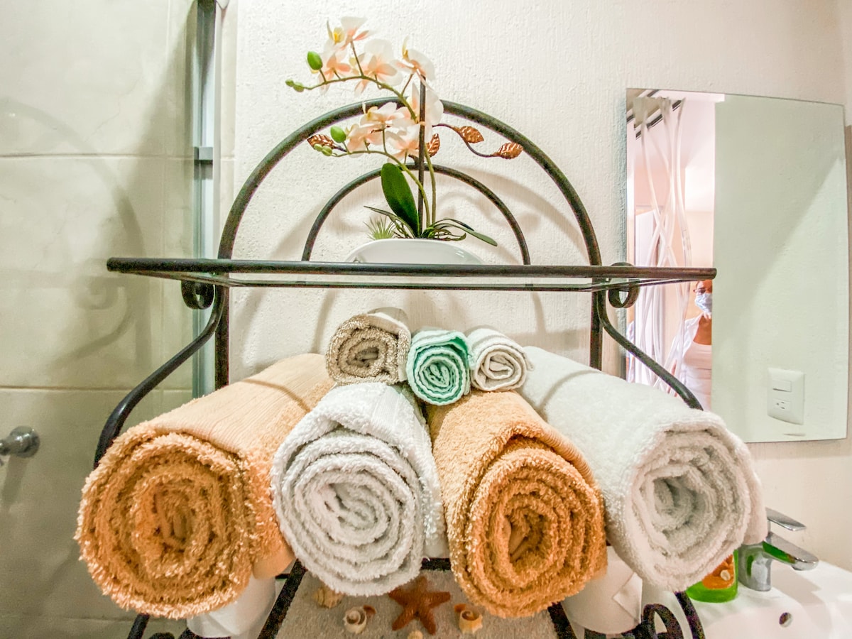 A neatly arranged towel rack displays an assortment of rolled towels in shades of orange, gray, and green. A small white orchid sits on top, accompanied by a mirror reflecting a softly lit bathroom space in the background.