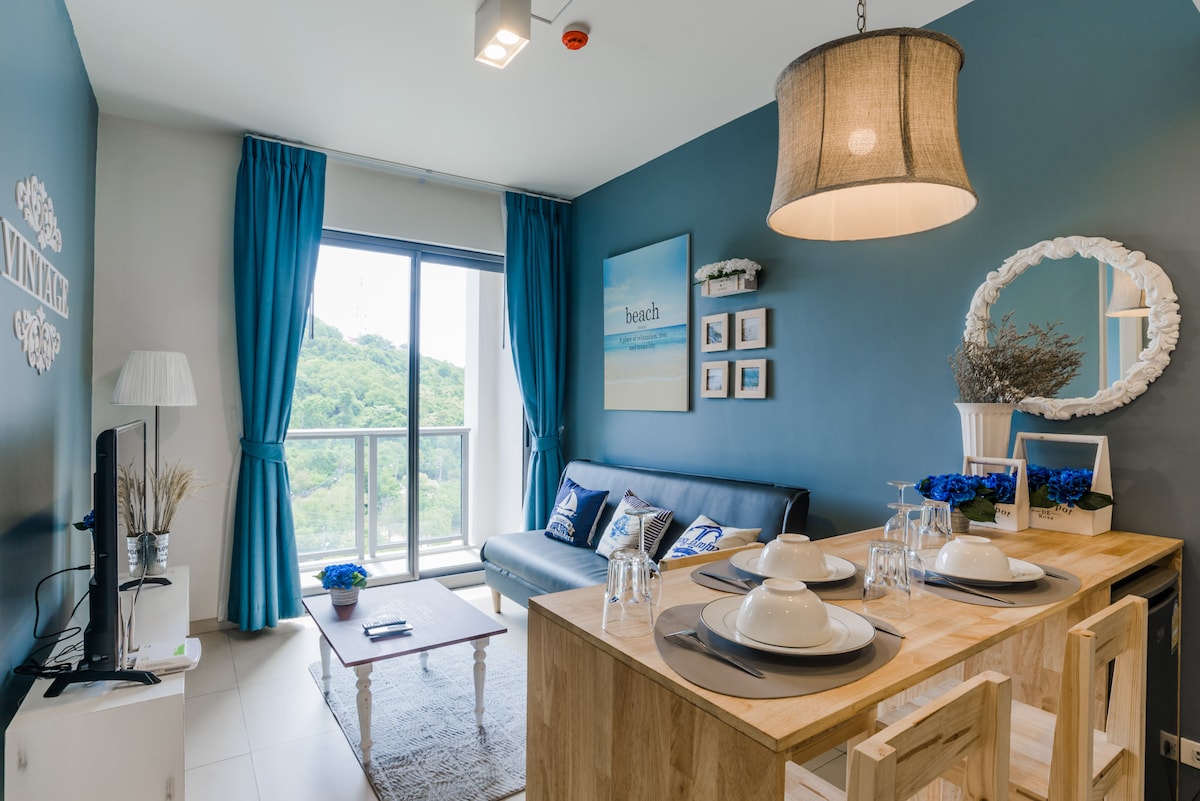 A cozy living area showcases a dark blue accent wall, complemented by matching curtains. A comfortable sofa is paired with a wooden dining table set for four. Natural light fills the space from the large balcony door, offering views of the surrounding greenery.