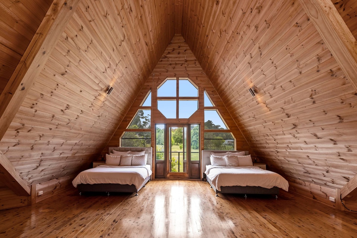 An architecturally designed bedroom features two spacious beds positioned under a high, sloped wooden ceiling. Large windows provide ample natural light and scenic views of the surrounding landscape. The warm wood tones enhance the inviting atmosphere of the room.