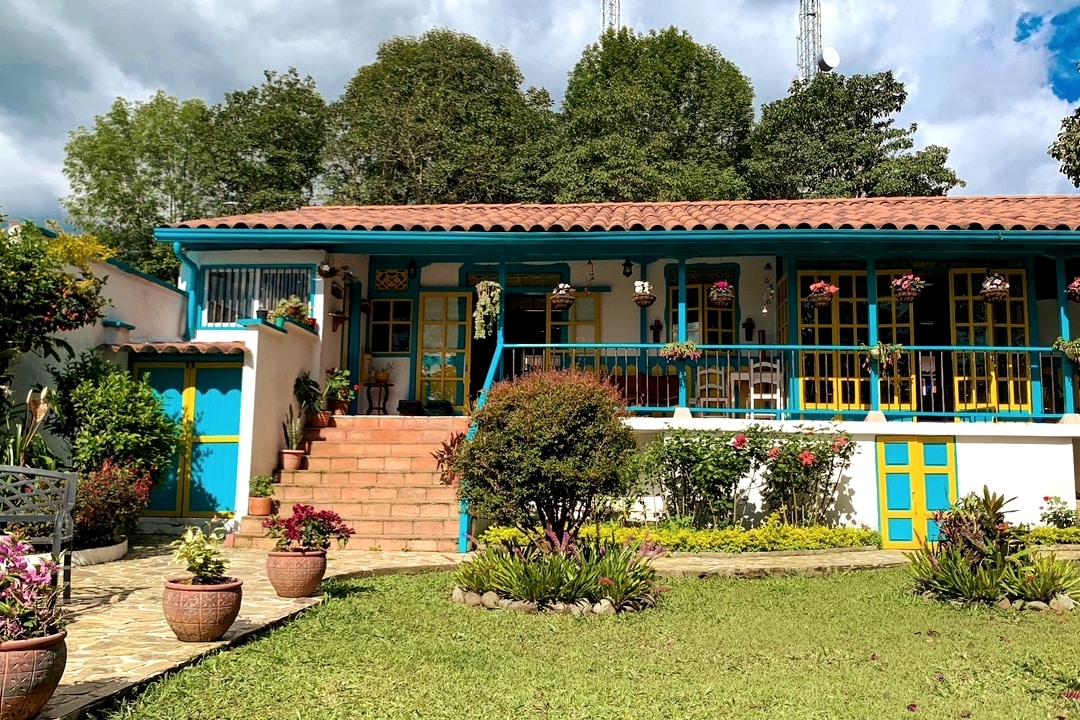 A traditional colonial-style house displays vibrant blue accents against white walls. The front porch features plants and hanging flower pots, while stone steps lead to the entrance. A well-maintained garden with colorful flora surrounds the home, creating a welcoming outdoor space.