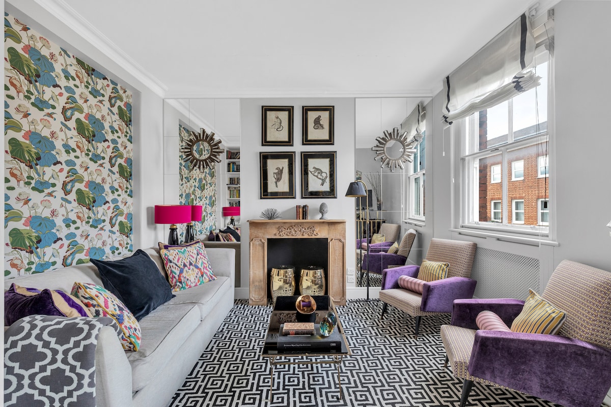 A modern living room features a light-colored sofa adorned with colorful cushions. Two upholstered chairs in vibrant purple complement the grey and white patterned rug. A mantelpiece with a mirror above stands against one wall, while decorative wall art adds character to the space.