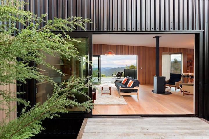 Modern open-plan living space with large sliding glass doors, cozy seating, and a view of mountains, framed by greenery