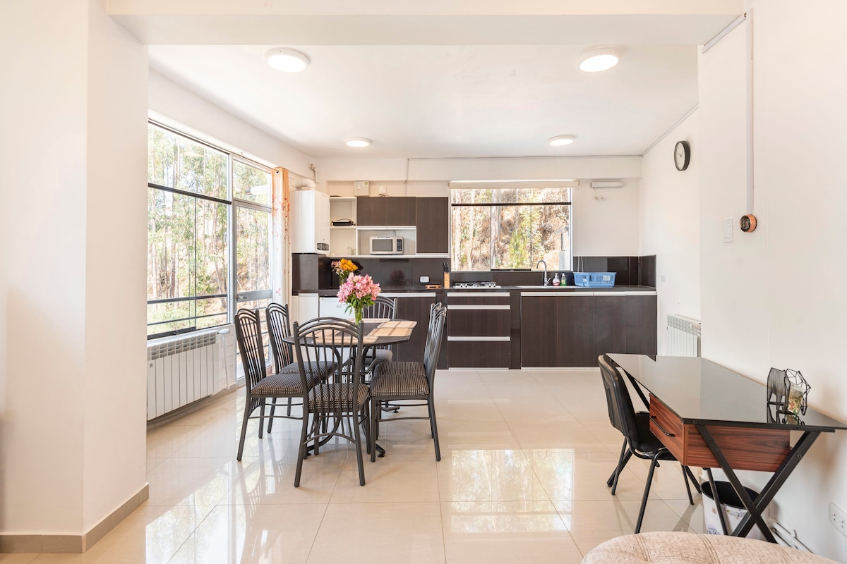 An open-concept living area showcases a dining table with six black chairs and a stylish table. A well-equipped kitchen is visible in the background, featuring modern appliances and dark cabinetry. Large windows allow natural light to fill the space, while greenery can be seen outside.