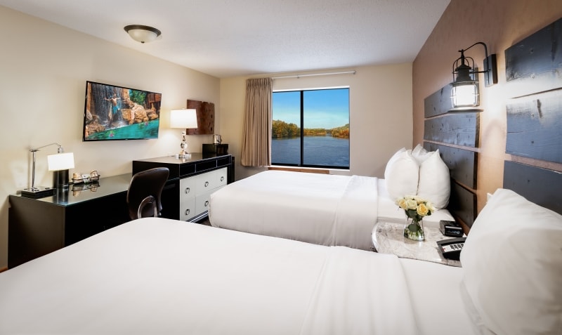 A well-lit suite features two spacious beds adorned with crisp white linens. A sleek desk is positioned nearby in front of a large window showcasing stunning views of the river. Soft natural light filters through sheer curtains, enhancing the calming interior.