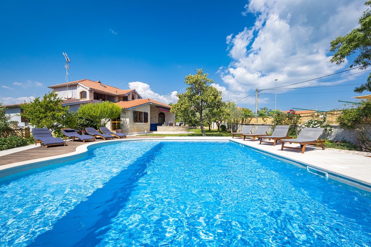 A spacious swimming pool is surrounded by lounge chairs under a clear blue sky. The pool is framed by a well-maintained garden with trees and shrubs. In the background, the holiday home features a rustic architectural design, complemented by various outdoor areas for relaxation.