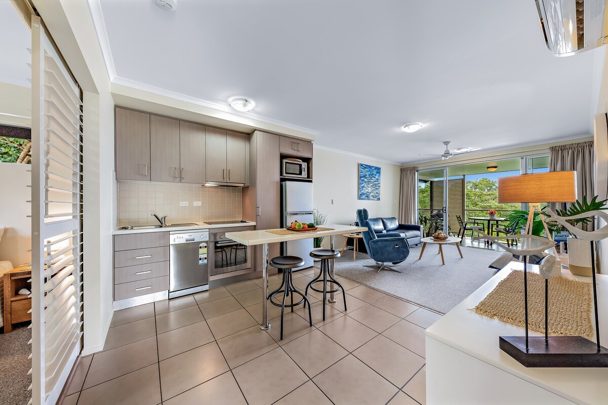 A modern kitchen and dining area is depicted, featuring sleek cabinetry and stainless steel appliances. A breakfast bar with seating is positioned next to a dining table. Light enters from large sliding doors leading to a balcony, overlooking lush greenery.
