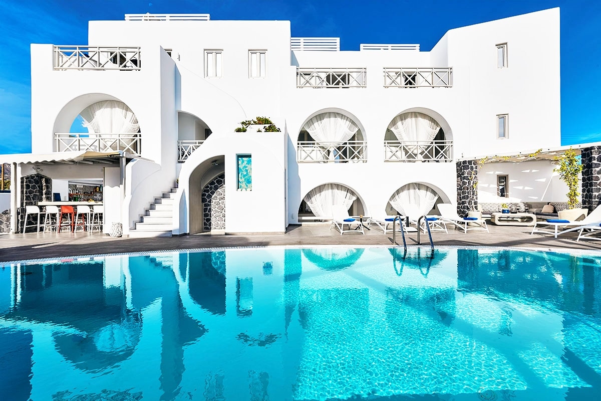 A modern white building features arched balconies overlooking a clear turquoise pool. The poolside area is adorned with sun loungers and a bar with high stools. Steps lead up to the building, surrounded by vibrant greenery, under a bright blue sky.