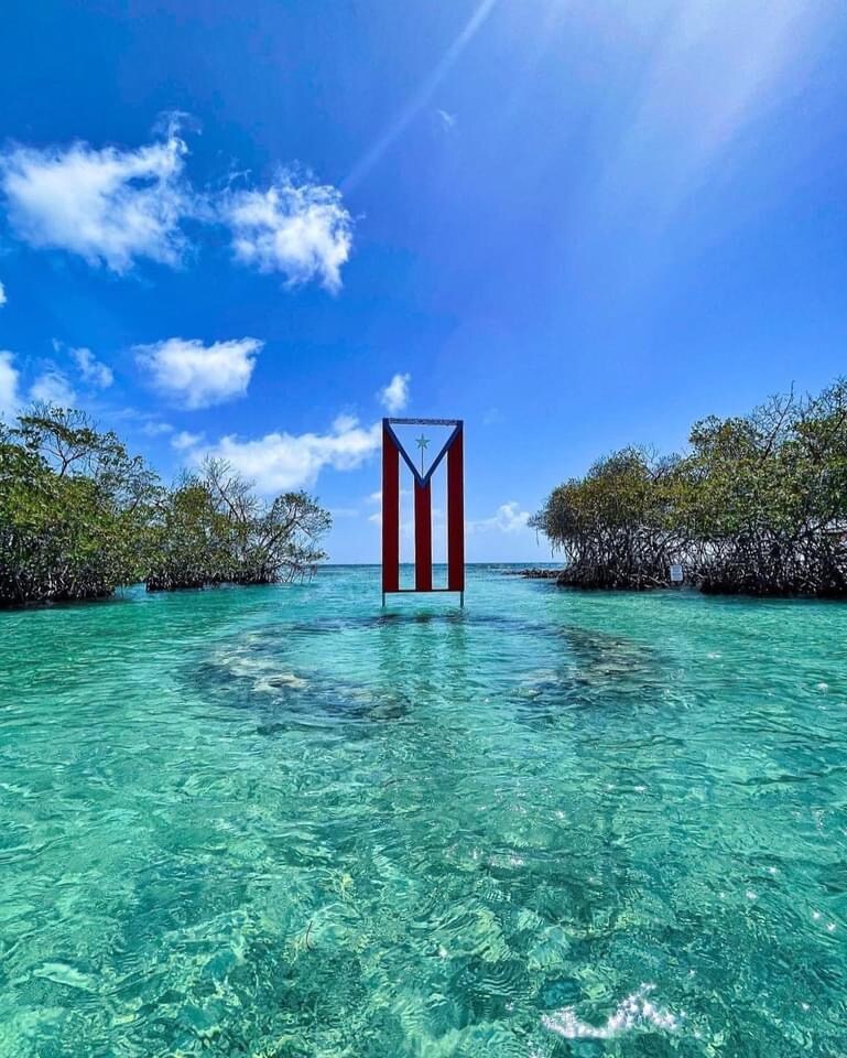 A striking red and white structure stands tall in the center of crystal-clear water, surrounded by lush green mangroves. The vibrant colors contrast against the bright blue sky, filled with fluffy white clouds, creating a serene landscape perfect for relaxation and exploration.