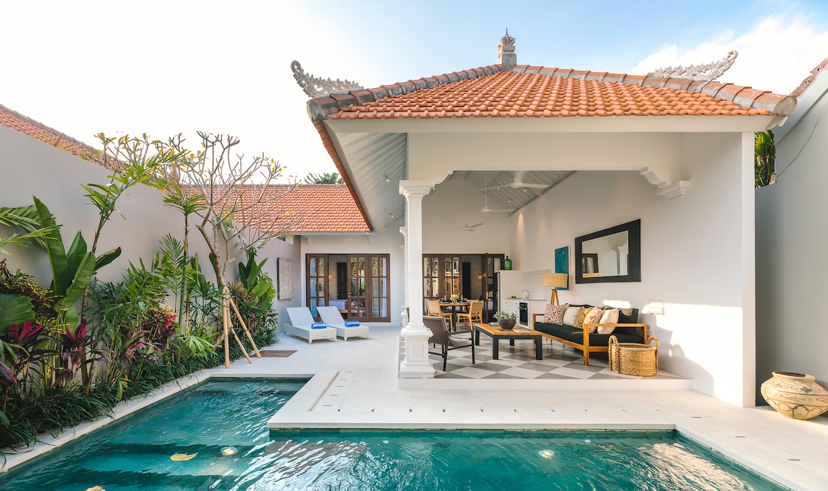 An open-air living area is surrounded by vibrant tropical greenery, featuring a sleek seating arrangement. A private pool is visible, reflecting the blue sky, while sun loungers offer a space for relaxation. The villa's contemporary architecture enhances the spacious and airy atmosphere.
