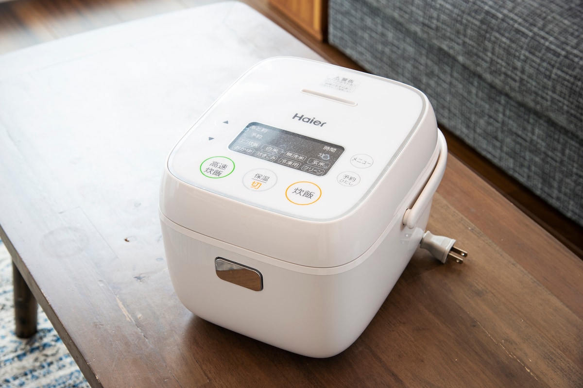 A white rice cooker is placed on a wooden table, showcasing a digital control panel with various buttons for settings. The cord is visible, providing power. The background features a soft, textured couch, suggesting a comfortable space for meal preparation.