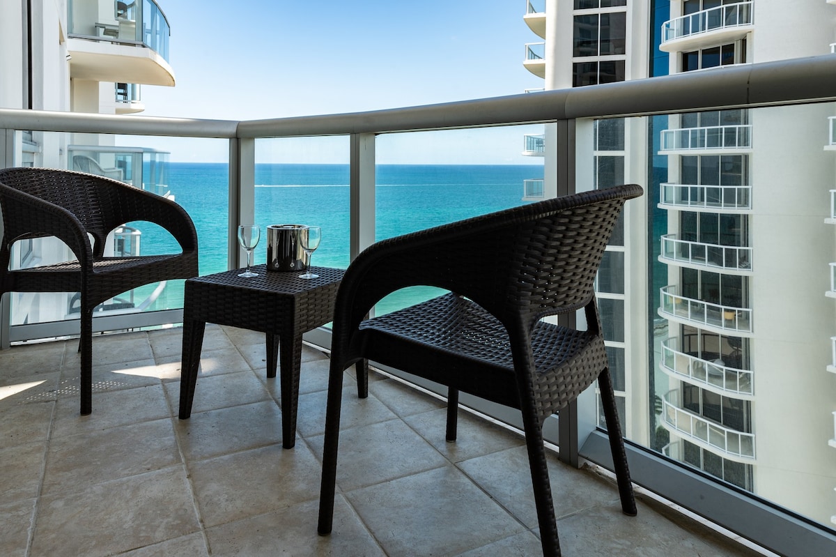 A balcony setup is featured with two woven chairs and a small table, providing a space for relaxation. The ocean view is prominently visible, showcasing clear blue waters and a tranquil atmosphere. Sliding glass doors facilitate access to the indoor area.