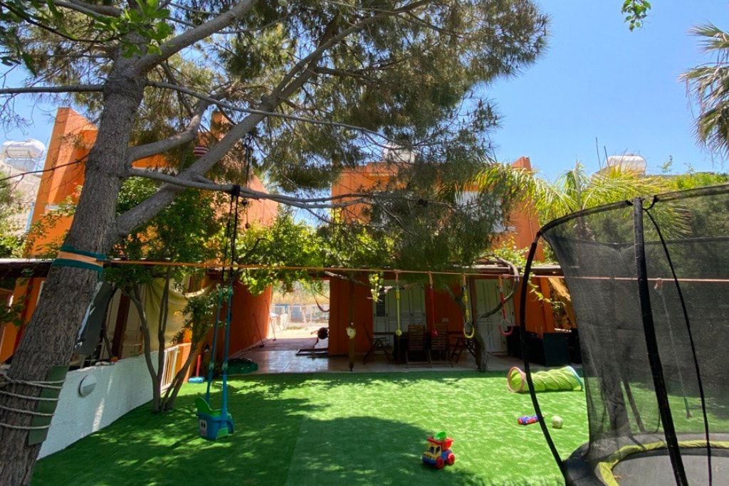 An outdoor area features a well-maintained green lawn surrounded by trees and shrubs. Two villas are visible in the background, their warm-colored exteriors contrasting with the vibrant greens. A trampoline and various toys are scattered across the lawn, indicating a family-friendly environment.