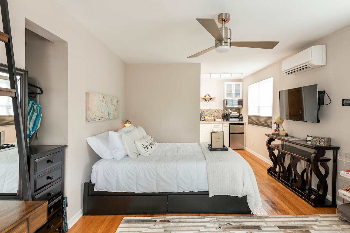 A spacious studio features a comfortable bed, accented with decorative pillows. A well-equipped kitchen is visible in the background, with modern appliances and ample counter space. Natural light enters through a nearby window, illuminating the hardwood flooring and minimalist decor.