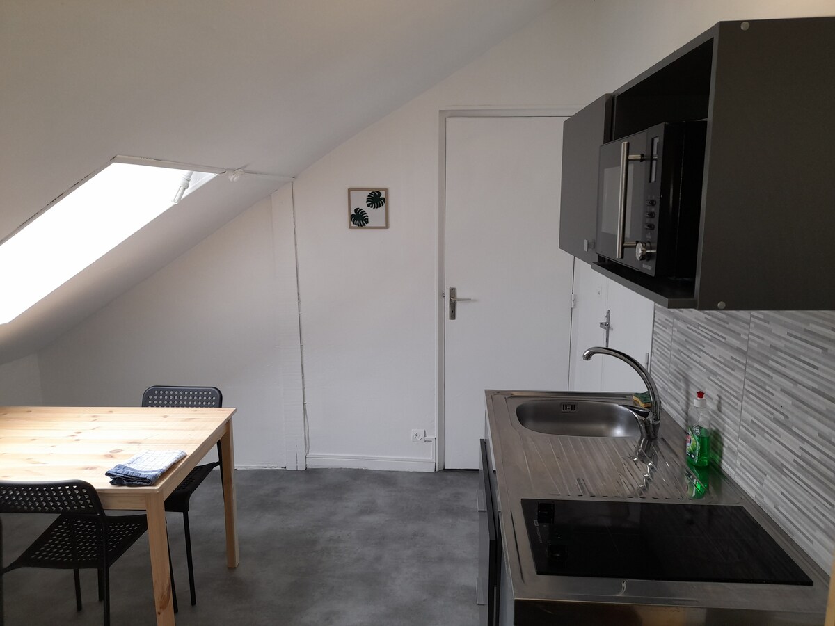 A functional kitchen area is presented with modern appliances and a sleek countertop. A dining table made of light wood is accompanied by black chairs. Natural light filters through a skylight, illuminating the space decorated with minimalistic wall art.