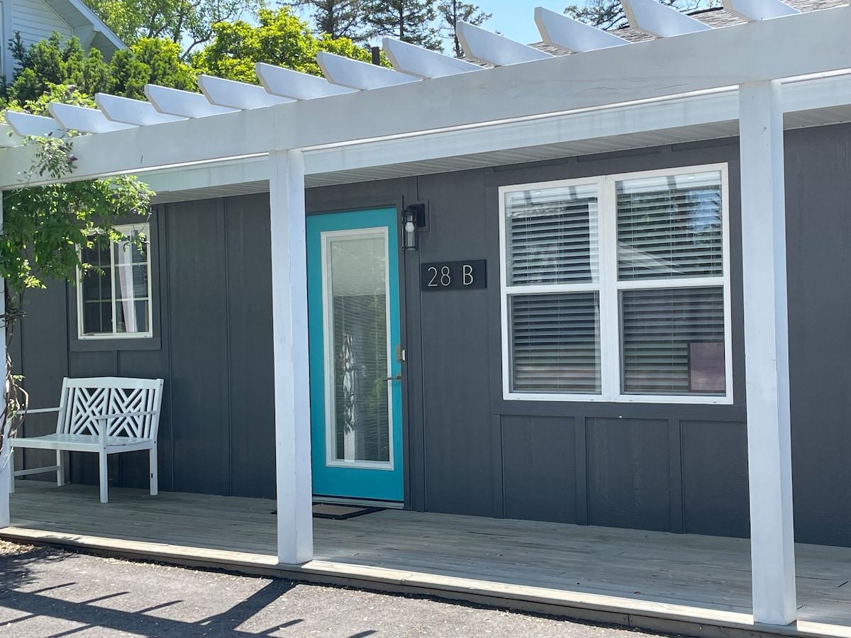 The entrance features a solid gray exterior enhanced by a bright turquoise door. White trim outlines the windows, while a covered porch is adorned with a white bench. Clean lines and a welcoming atmosphere characterize the overall design.
