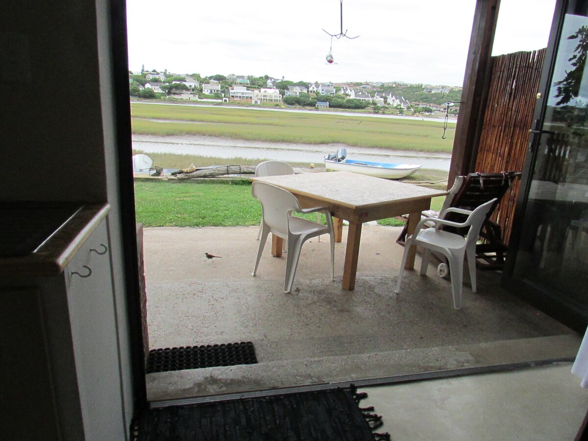 A simple outdoor dining space features a wooden table and four white chairs on a concrete patio. The river is visible beyond the lawn, with boats moored nearby. Natural light fills the area, providing a serene view of the surrounding landscape.