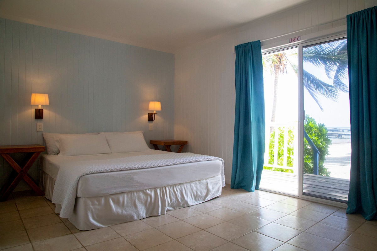 A spacious sleeping area features a large bed adorned with crisp white linens. Natural light filters through sliding glass doors, revealing a view of palm trees. Wooden side tables are positioned beside the bed, and light blue walls lend a calm ambiance.