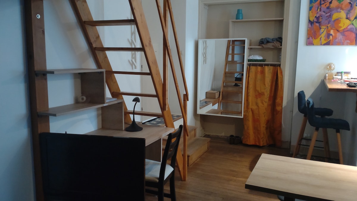 An interior view of the studio showcases a wooden ladder leading to the mezzanine with a queen size bed. A small dining table and chair are positioned beside a mirror and shelf, while a cozy seating area is visible to the left.