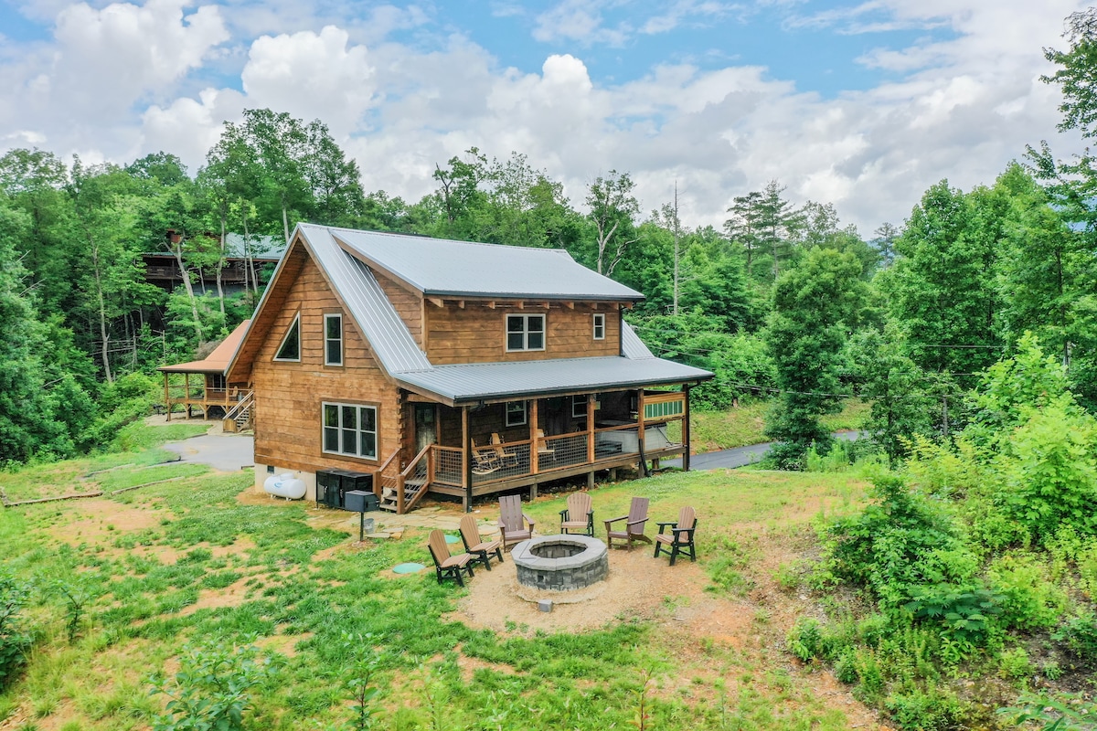 A spacious log cabin is surrounded by lush greenery, featuring a front porch and an outdoor fire pit with seating arranged around it. The roof displays metal accents, while the natural landscape enhances the peaceful setting.