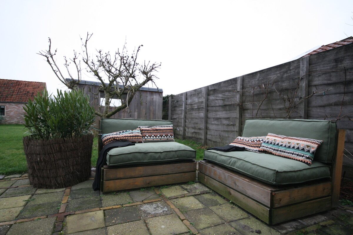 Two wooden lounge chairs with cushions, positioned in a small patio area. A decorative plant is seen nearby, with a backdrop of a weathered wooden fence. The patio features stone tiles and is surrounded by grassy areas.