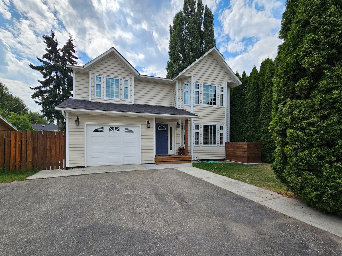 A two-story house is shown, featuring a light-colored exterior with large windows. A driveway leads to a garage, and a well-maintained lawn is visible in front. Tall evergreen trees border the property, providing privacy. Cloudy skies create a serene backdrop.