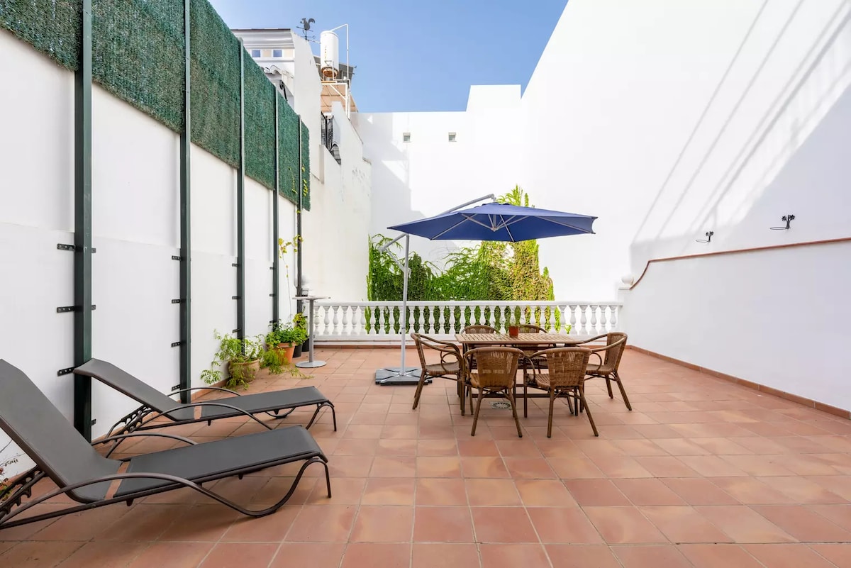 An outdoor terrace features a dining table surrounded by chairs and two loungers positioned for relaxation. A large umbrella provides shade over the dining area, while green plants add a touch of nature against the white walls and bright blue sky.