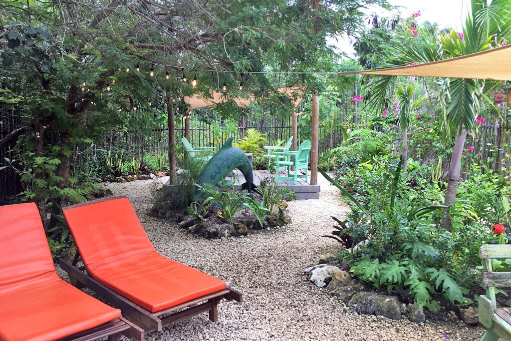 A tranquil garden area features orange lounge chairs positioned on gravel, surrounded by lush greenery and vibrant flowers. A wooden gazebo holds a light green table and chairs, providing a cozy gathering spot beneath a canopy of shade from surrounding trees.