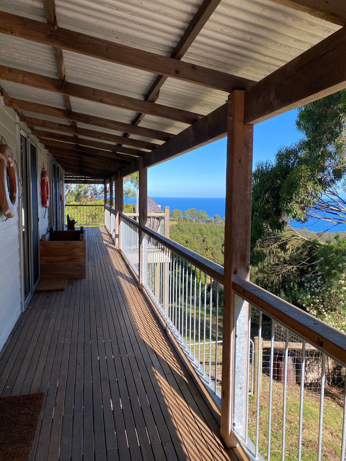 A wooden deck is viewed from an angle, lined with railings and offering expansive views of the ocean in the distance. The space is illuminated by natural light, and nearby trees add to the tranquil atmosphere.