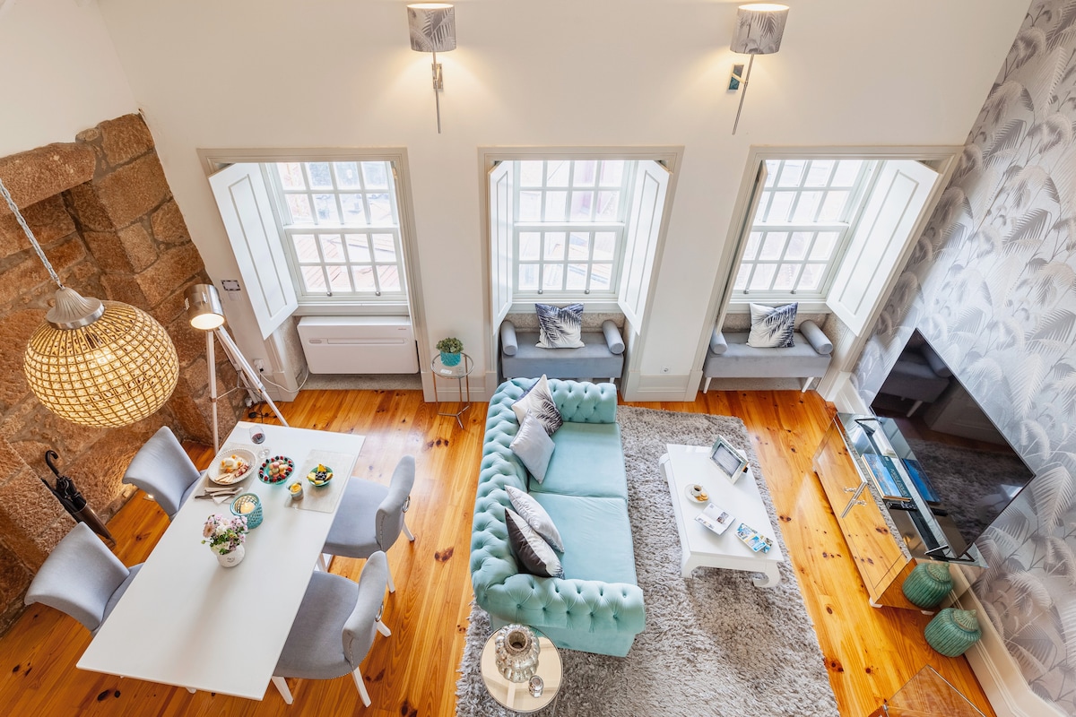 An open living area showcases a light blue sofa with decorative cushions and a rug beneath. There is a dining table with four chairs, accompanied by a variety of snack items. Large windows provide abundant natural light, framed by elegant white shutters.