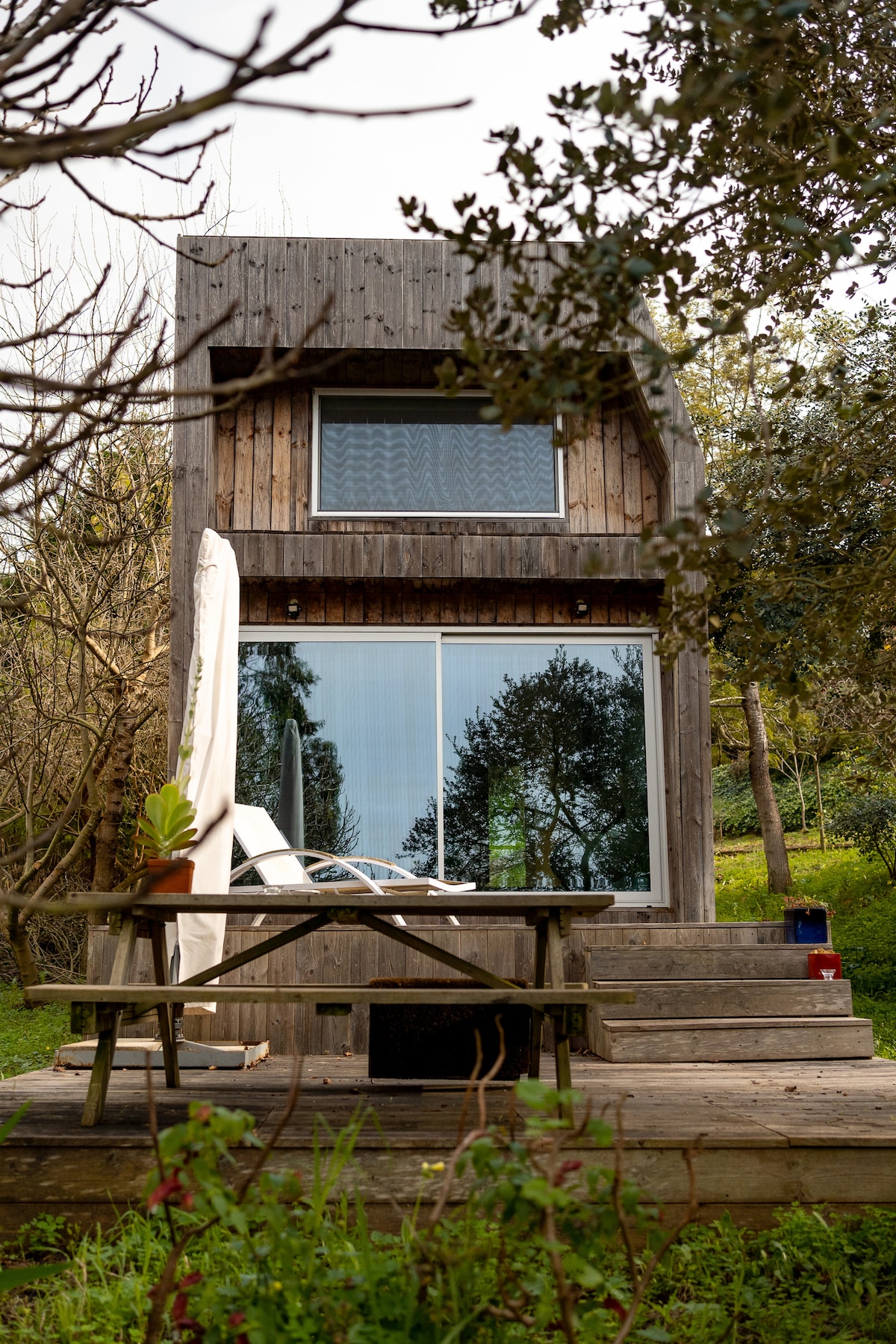 A modern wooden house features a sloped roof and expansive glass windows, providing a view of the surrounding greenery. A small wooden patio is visible in the foreground, complemented by a picnic table with seating, creating an inviting outdoor space.