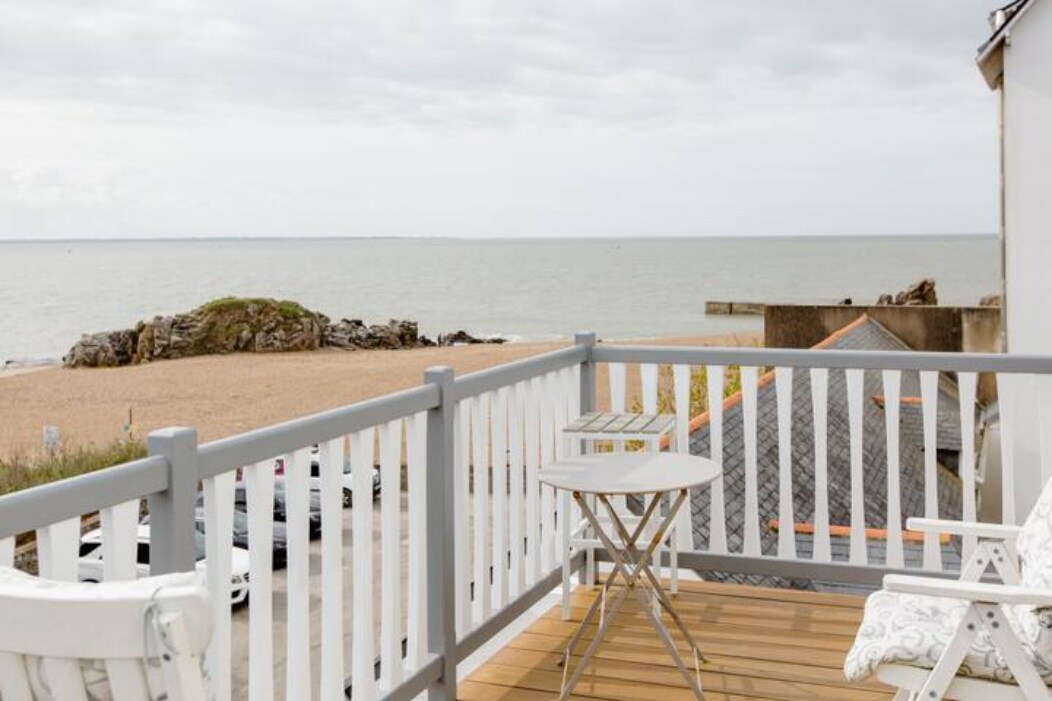 A balcony adorned with white wooden railings showcases a small round table and two chairs, offering views of the beach and the sea beyond. The coastline is visible in the distance, accompanied by scattered rocks and a sandy shore.
