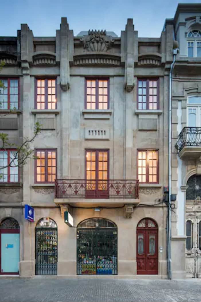 An elegant historic building features a symmetrical façade with intricate architectural details. Large windows, adorned with red frames, allow warm light to escape. A balcony above the ground floor is framed by decorative stonework, adding to the character of the structure.