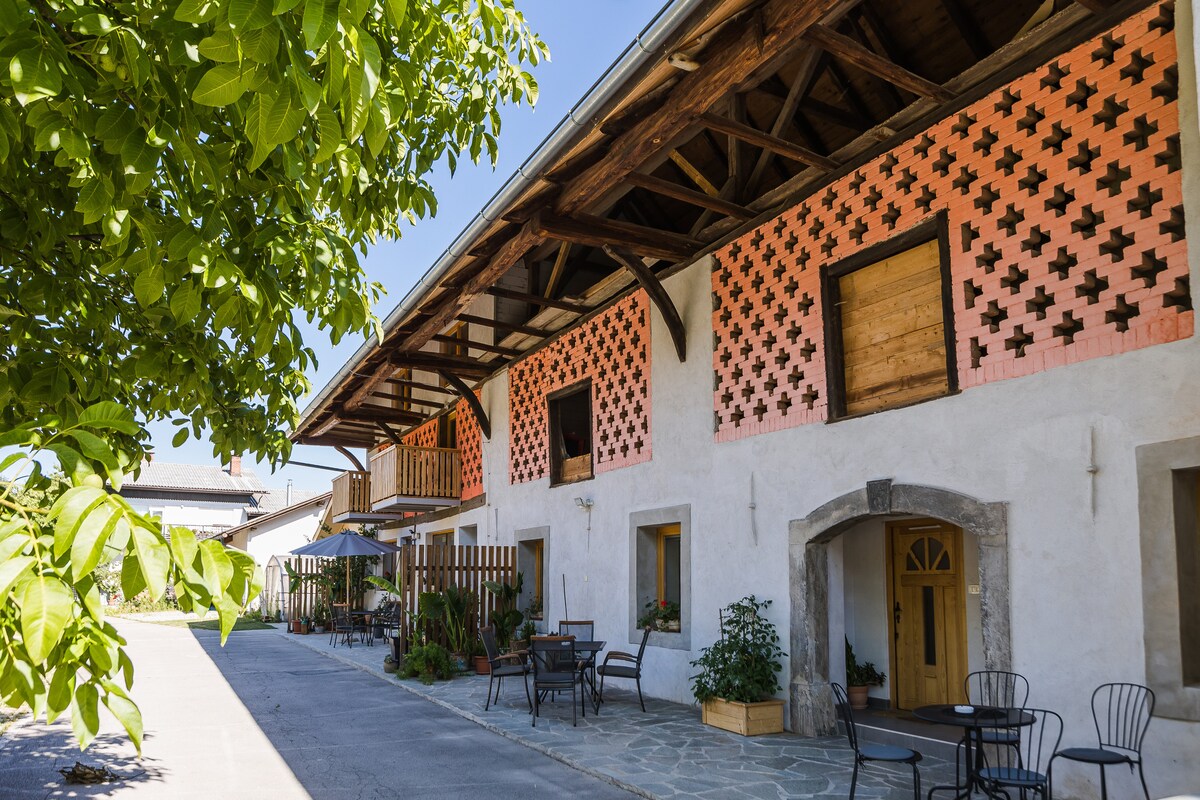 The exterior of the historical barn showcases a charming blend of traditional architecture and modern design. Orange brick patterns accent the façade, while wooden balconies add character. A spacious courtyard is visible, featuring outdoor seating arrangements and greenery that enhances the tranquil environment.