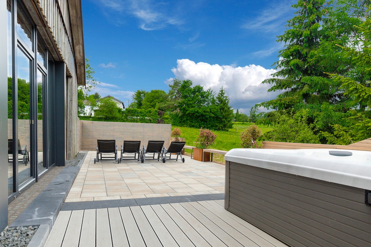 A spacious terrace features a hot tub alongside comfortable lounge chairs, inviting relaxation. Lush greenery and trees are visible in the background, complementing the serene outdoor setting. The area is well-maintained and designed for guests to enjoy the natural surroundings.