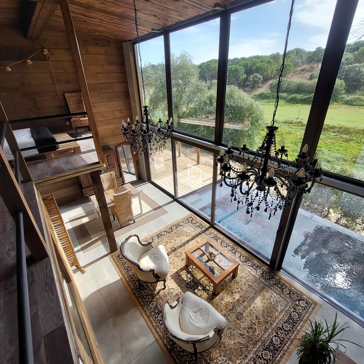A spacious living area showcases large glass windows that provide views of the surrounding greenery. Elegant chandeliers hang above, while two cozy armchairs face a wooden coffee table. A decorative rug adds warmth to the space, enhancing the connection with nature outside.