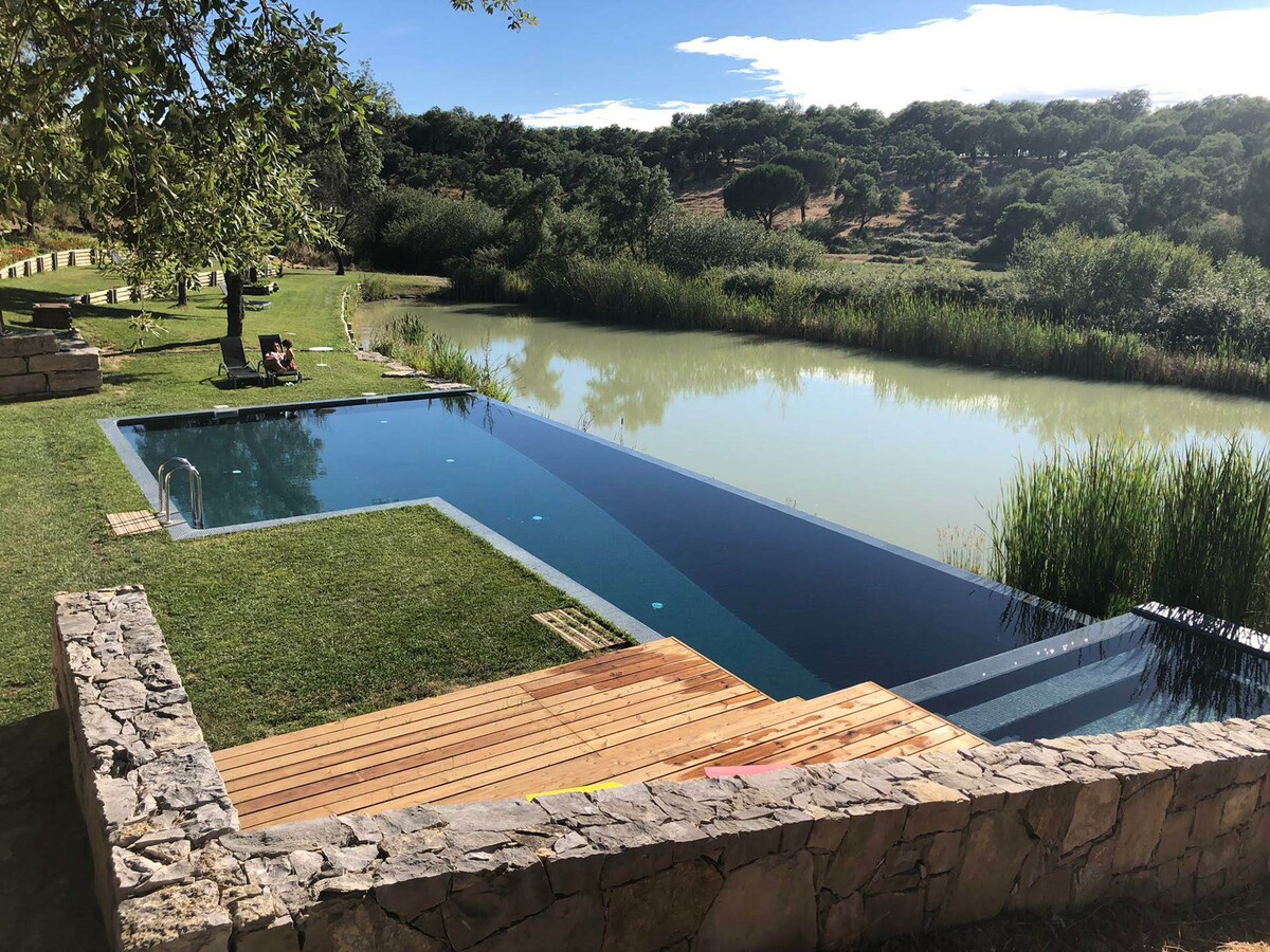 An infinity pool is positioned against a picturesque natural backdrop, featuring a smooth transition from wooden deck to the water. Lush greenery surrounds the pool area, with a serene lake visible nearby, enhancing the peaceful atmosphere of the setting.