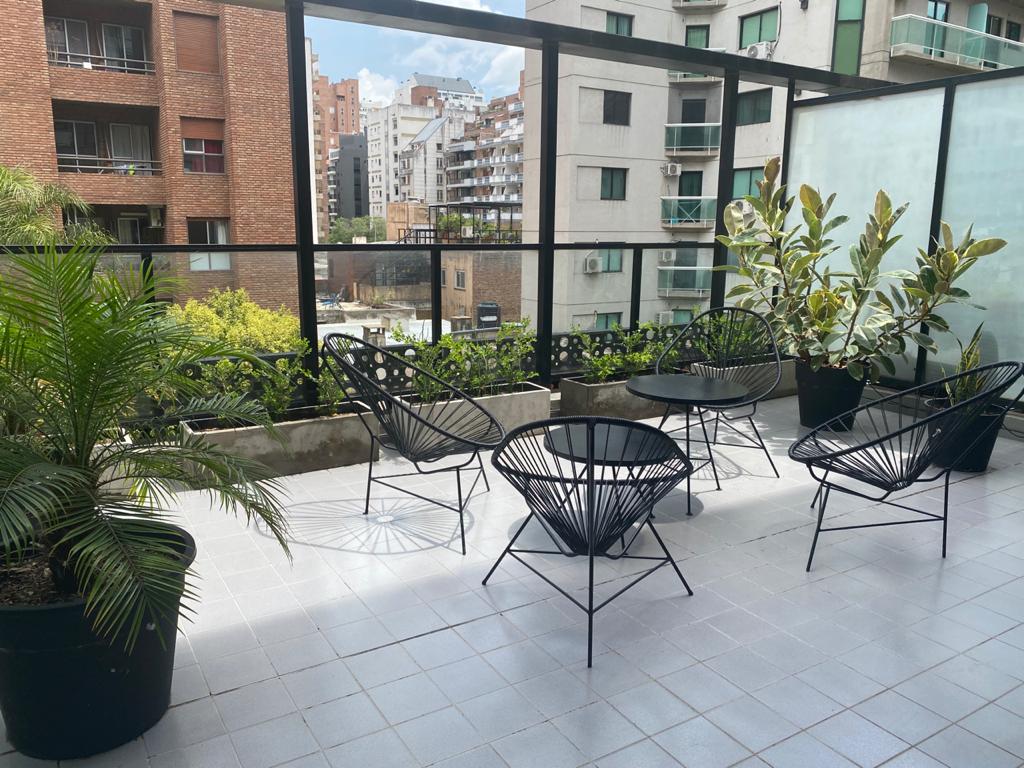 A private terrace is featured, adorned with modern black chairs and small circular tables. Green plants provide a touch of nature, while the surrounding buildings offer an urban backdrop. The area is bathed in natural light, creating a relaxed outdoor space.
