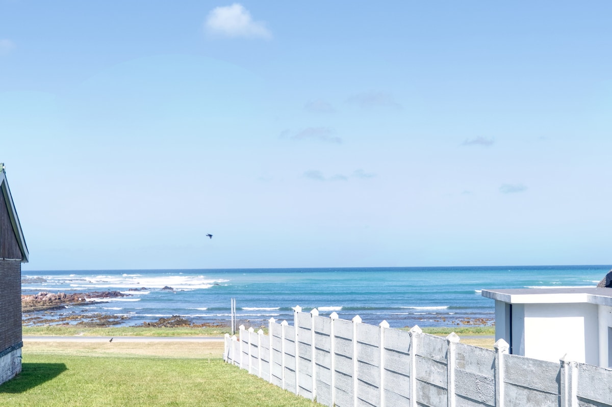 A vast view of the sea is presented, featuring gentle waves under a clear blue sky. A neatly maintained grassy area in the foreground is bordered by a light-colored fence, contrasting the natural landscape. The serene atmosphere is enhanced by a distant rocky shoreline.