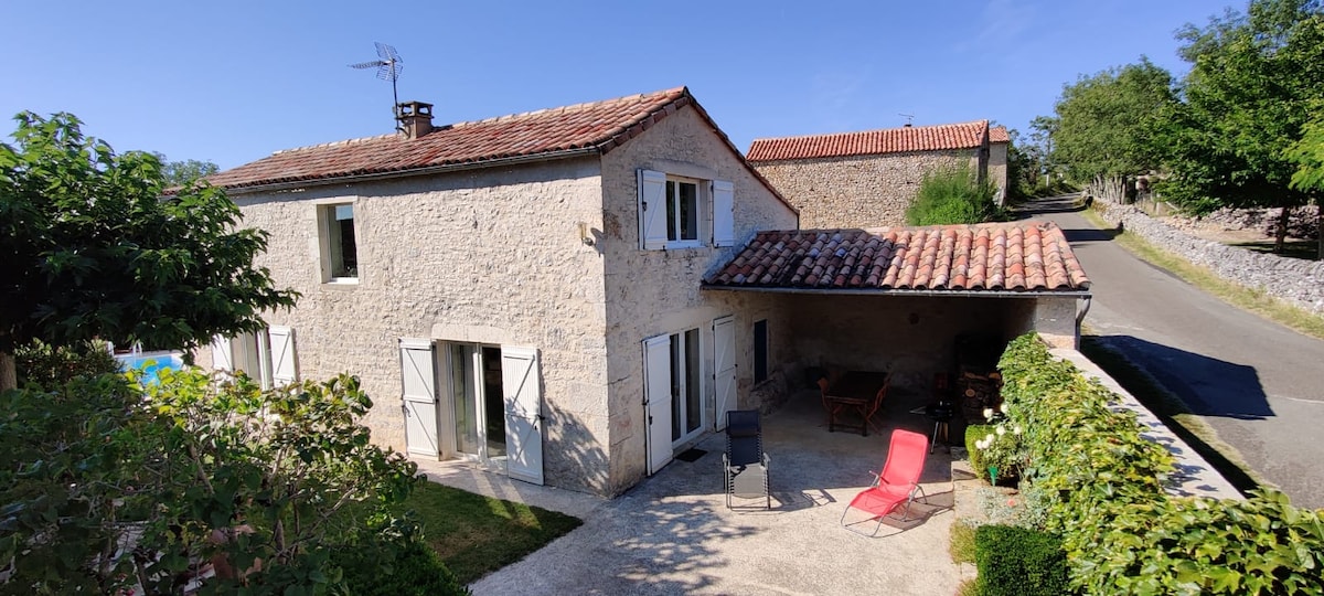 A stone house with a red-tiled roof is featured, complemented by white shutters. A shaded patio area is visible, equipped with outdoor seating, while greenery surrounds the home. A quiet road leads past the property, enhancing the serene setting.