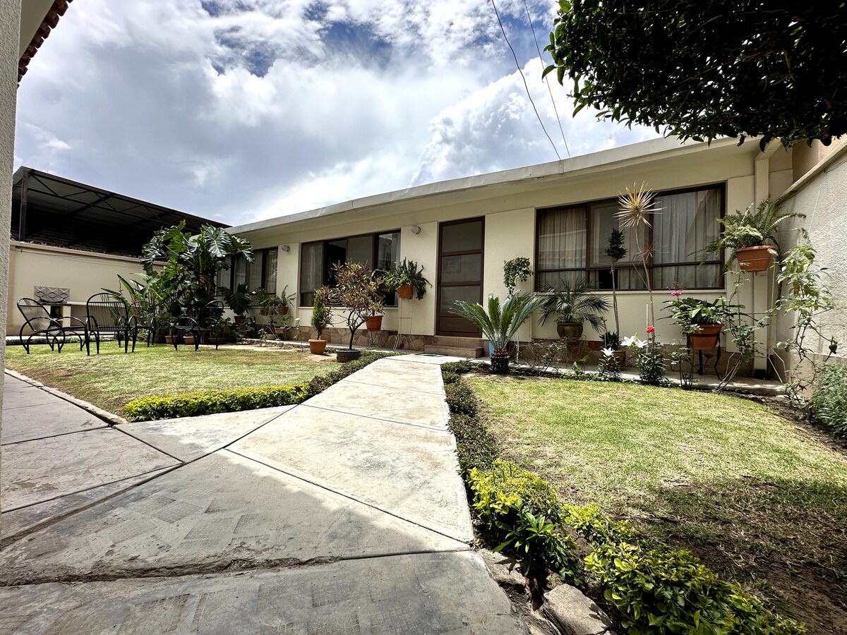 A charming garden pathway leads to the entrance of a single-story apartment surrounded by vibrant greenery. Potted plants and small shrubs enhance the outdoor space, complemented by a seating area equipped with chairs. Natural light brightens the facade, creating a welcoming atmosphere.