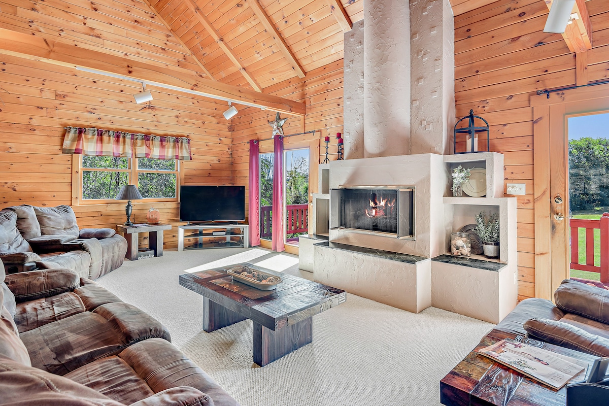 A spacious great room features a wooden ceiling with exposed beams and a cozy fireplace as its focal point. Comfortable seating is arranged around a wooden coffee table. Large windows allow natural light to flood the space, offering views of the surrounding greenery.
