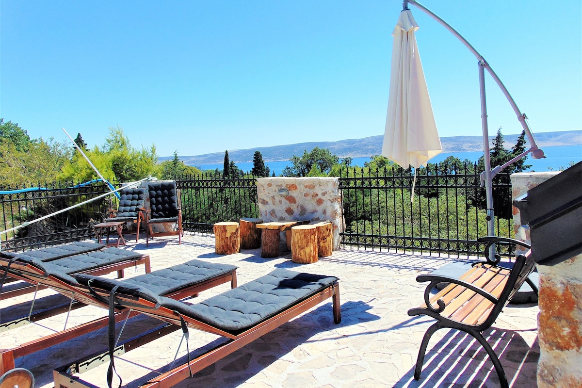 A spacious outdoor terrace is equipped with several loungers and a dining area composed of natural wooden stools. A large umbrella offers shade, while the area is surrounded by a stone railing, providing views of the lush landscape and distant sea.