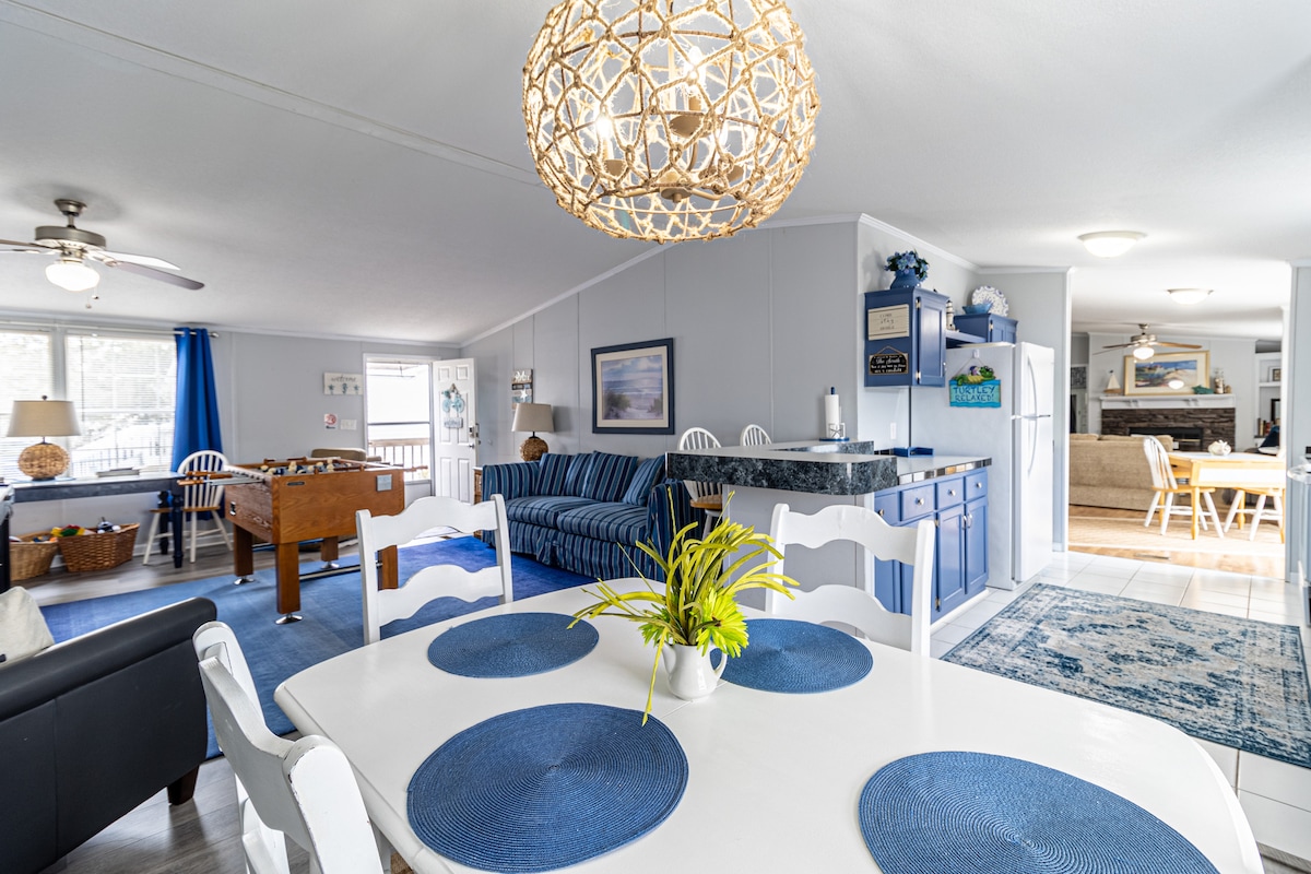 An open living space showcases a dining area with a white table set for six, accented by blue placemats. A large chandelier illuminates the room, which features a cozy sitting area with blue-striped sofas. The kitchen area is visible in the background, creating a welcoming atmosphere.