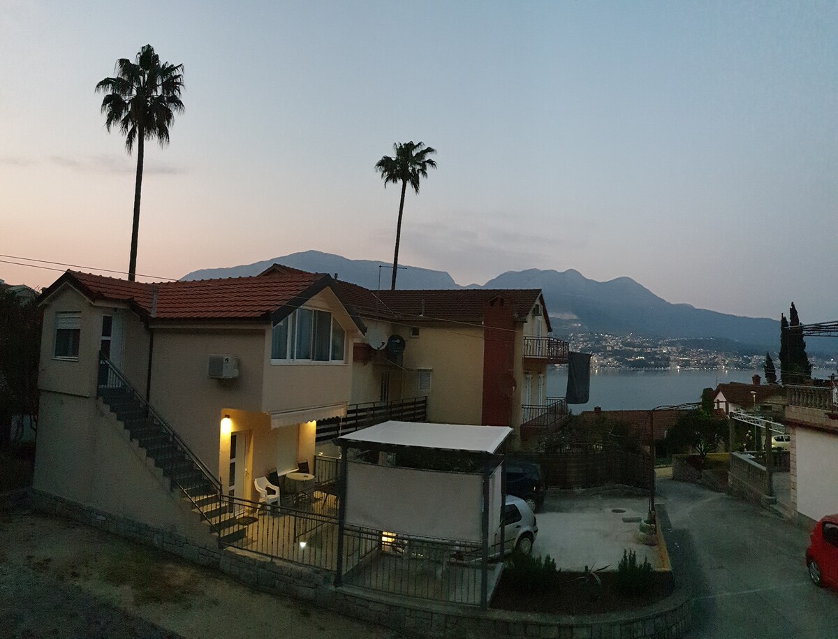 An exterior view of the two-story building reveals a serene setting at dusk, framed by palm trees. Soft lighting highlights the entrance, while mountains and a coastal view are visible in the background, creating a tranquil atmosphere.