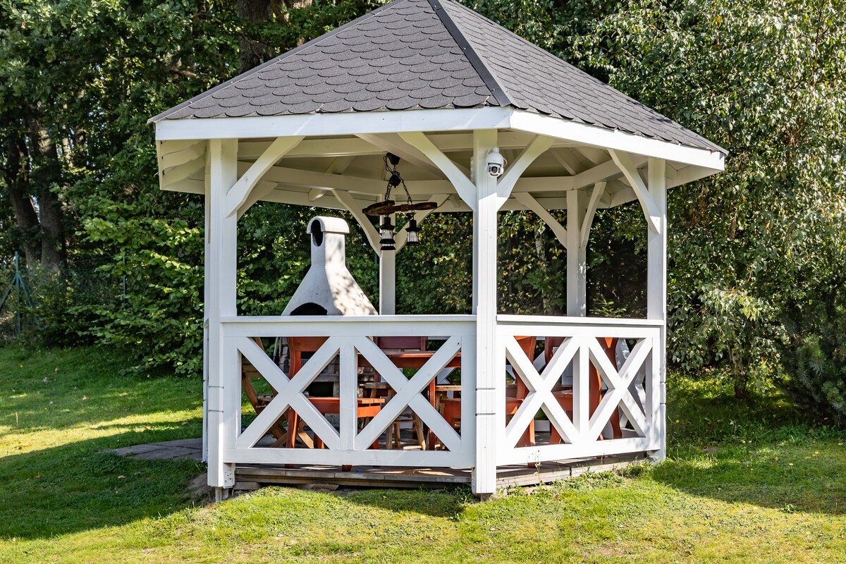 A spacious wooden gazebo with an inviting, open design is featured. It includes a central fireplace and is surrounded by a manicured lawn. Wooden tables and chairs are arranged inside, providing a comfortable space for gatherings in a natural setting.