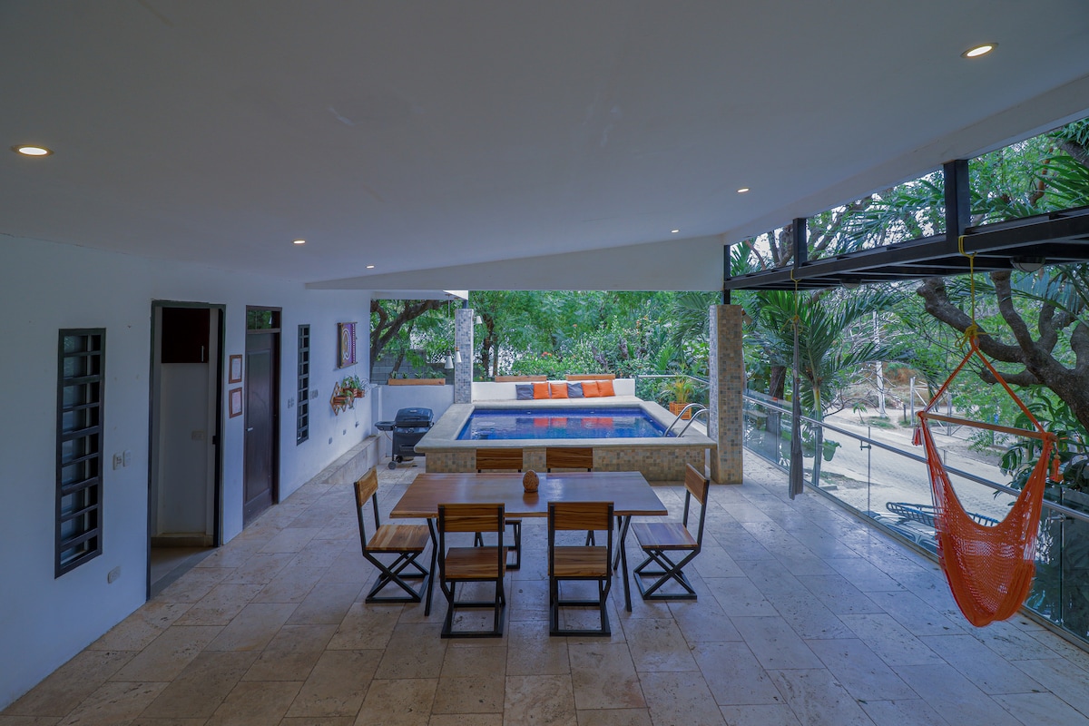An open-plan living area features a dining table with wooden chairs. A plunge pool can be seen through large glass windows, surrounded by lush greenery. A vibrant orange hammock adds a touch of color to the space, providing a cozy spot for relaxation.